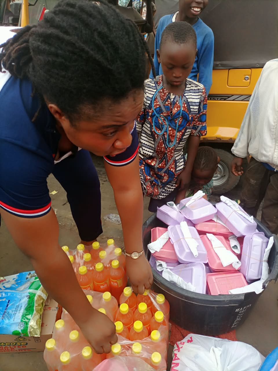 A volunteer unpacking crates of drinks beside packed meals as children look on