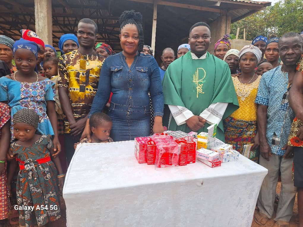 Boxes of donated medicines laid out on a table at a rural health outreach, with Nurse Vivian Okafor and the community behind them